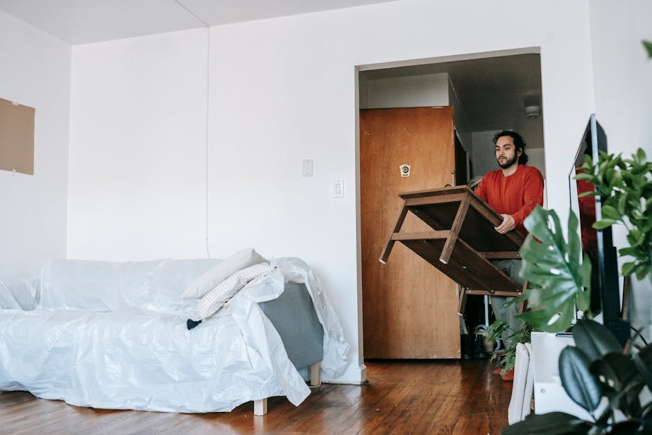 A male mover is seen inside a residential room holding a wooden table while standing near an open doorway, preparing to load it onto a moving vehicle. The room features a white sofa covered with a pla
