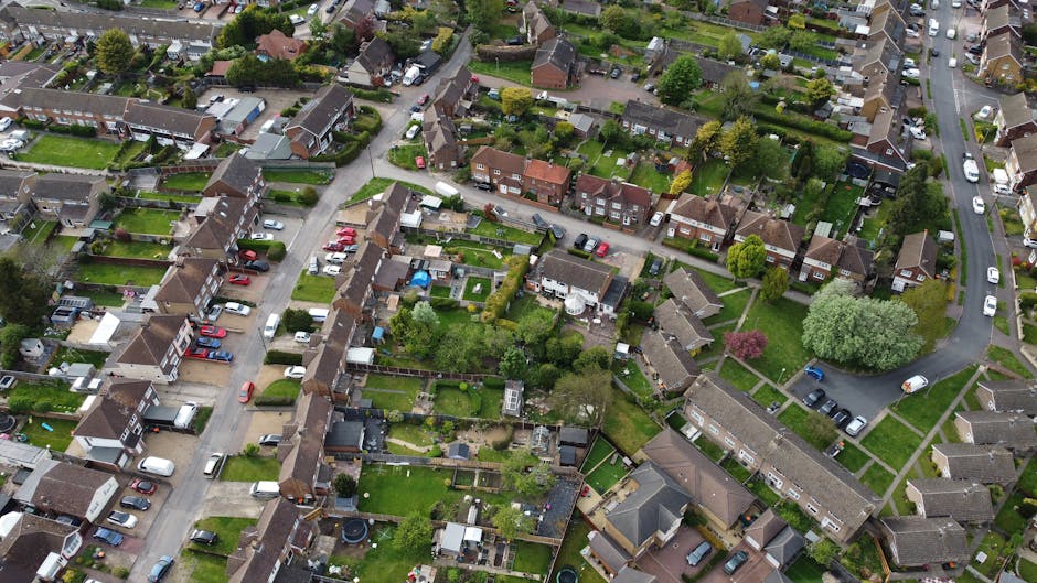 An aerial view of a residential street showing a row of terraced houses with small back gardens. The gardens vary in size and features, with some having grass lawns, paved patios, sheds, or garden fur