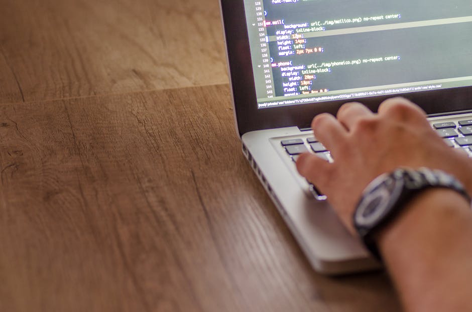 A person's hand wearing a wristwatch operates a laptop with a silver body on a wooden surface, displaying lines of code on the screen. The background and environment are not visible, focusing solely o